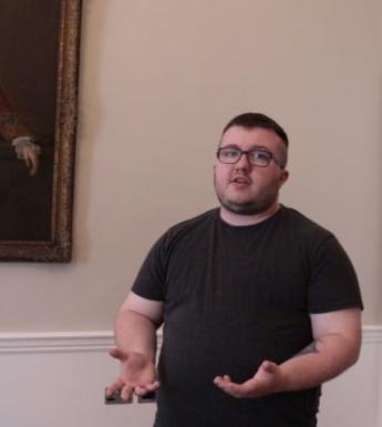 Video still of a young trans man standing in front of a gilt-framed portrait in a room with pale walls and a white door.