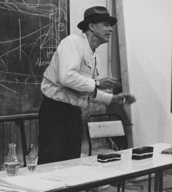 A black and white photograph of Joseph Beuys delivering a lecture at the Crawford Art Gallery. He is animated, in the process of speaking. Behind him is a blackboard which is covered in diagrams and writing.