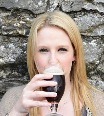 A woman with long blonde hair stands against a stone wall drinking a glass of stout