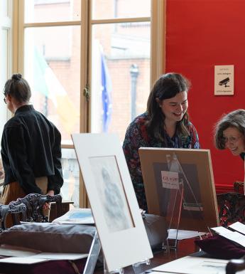 Three female figures in a red room, looking at a collection of drawings, books and other items that have been spread on a table. 