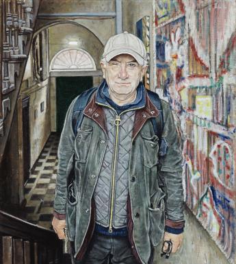 A man stands in a stairwell. Behind, we see a door with a fanlight and a tiled hallway. To his left, a colourful painting hangs on the wall. He is wearing jeans, a baseball cap, a wax jacket and a puffer jacket, and has a backpack on his back.