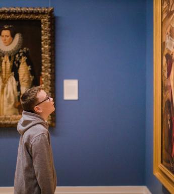 Boy wearing glasses looking at a large gilt-framed painting in a Gallery