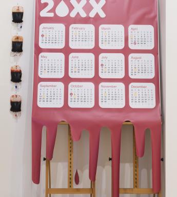 Installation shot of a large red calendar, mimicking blood dripping, mounted on an easel, with four blood bags hanging on the wall beside it