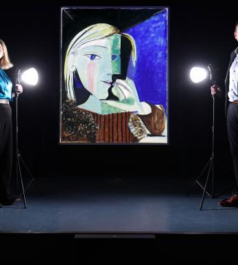 Two figures stand in semi darkness on either side of a large painting by Picasso. In front of both of them is a spotlight.