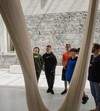 A Gallery guide speaks to a group of school students in front of a wooden sculpture