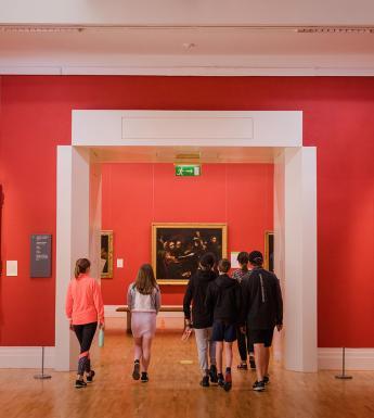 A group of young people walking away through an open doorway in an art gallery with red walls and gilt-framed oil paintings