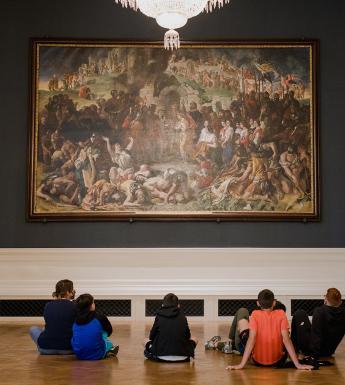 School students sit on floor in front of a large oil painting as a woman wearing a face mask speaks to them