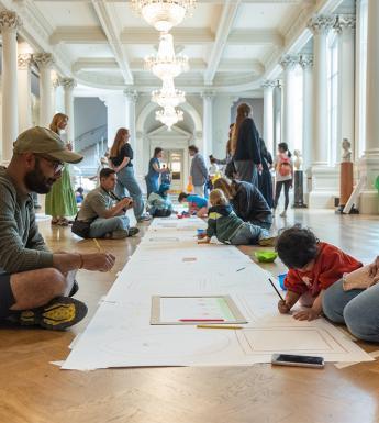 A family draw pictures on a long sheet of paper, which is laid on the floor of a grand room.