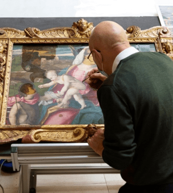 A man in a green jumper leans over an oil painting in a gilt frame with a small brush.