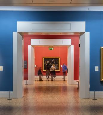 Photograph of a group of people looking at a painting, taken from down a long red and blue corridor.
