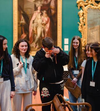 A teenage boy takes a photo on a bright orange camera in a grand gallery space. A group of five teenage girls watch on.