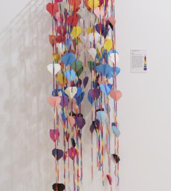 Installation shot of dozens of colourful origami hearts strung on strings and hanging down a white wall