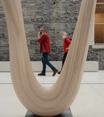 A man and woman wearing face masks walk past a wooden sculpture in the Gallery