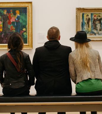 Photo of two women and a man seated on a bench, with backs to camera, looking at two paintings on a white wall.