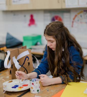 A young girl sits at a desk painting a cardboard and paper sculpture with brightly coloured paints