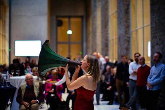 Photo of people attending an event with a woman playing speaking through a gramophone horn.