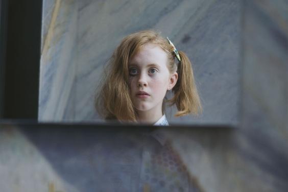 A photograph on paper. The head of a young girl is clearly visible, as though reflected in a square of mirror - on one side of her head her hair is pinned up in a ponytail, on the other side it is loose to her shoulders. One of her eyes has black markings under it. She is surrounded by abstract patterns - we can see her shirt faintly, as though reflected through a window. Out of focus beneath her face we see three brass fittings, like the fixtures of a bath.