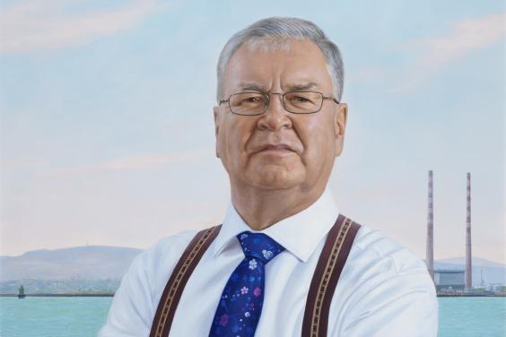 Three quarter length portrait of a man. He is wearing a white shirt, braces, and a blue tie. In the background we can see the sea, the mountains, and the Poolbeg towers.