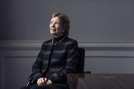 Former President of Ireland Mar Robinson sits beside a wooden table with her hands clasped in her lap. She is dressed in a grey and black striped jacket with a brooch at the lapel, and she gazes upwards and to her right.