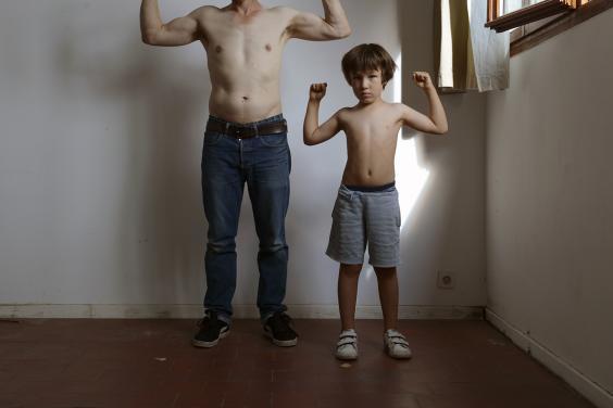 Two figures - an adult and a child - stand in an empty room against a white wall. Both are topless, with the adult figure in jeans and the child in shorts. They both have their arms raised as though holding an imaginary weight over their heads. The adult figures head is not visible, but the child looks directly at the camera, and there is a shaft of light from the open window behind him.  
