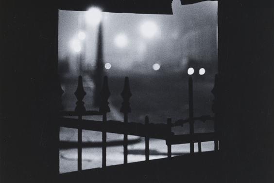 Black and white fine-art photograph of a gloomy city night scene viewed through a crack in a fence.