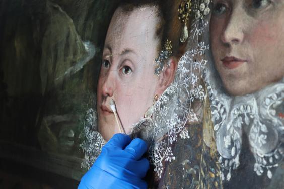 Close-up view of a blue-gloved hand wiping an oil painting of two women's faces.ing cotton swabs