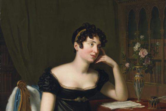 A woman sits at a desk, pen in hand, paper in front of her, lost in thought