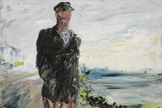A man in a dark overcoat with his hands in his pockets, wearing a cap. In the background, the sea.  