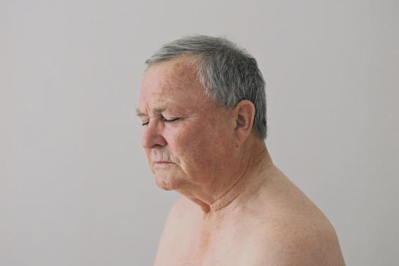 Photograph of an older man's head and shoulders with grey hair and his eyes closed sitting nude on a grey background