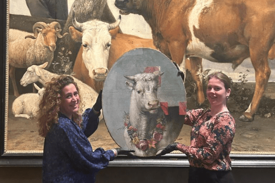 Two women hold up a small oval painting of a bull's head in front of a much larger painted scene featuring a bull
