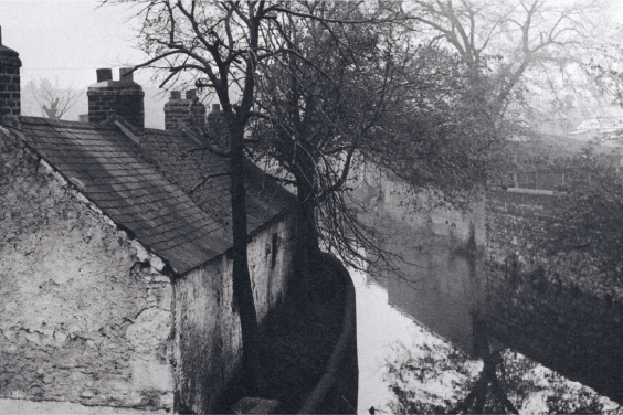 A black and white photograph of cottages beside a river. Bare trees beside the cottages are reflected in the river.
