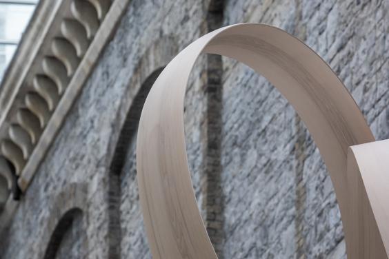 Cropped image of a curved grey wooden sculpture in front of a grey stone wall.