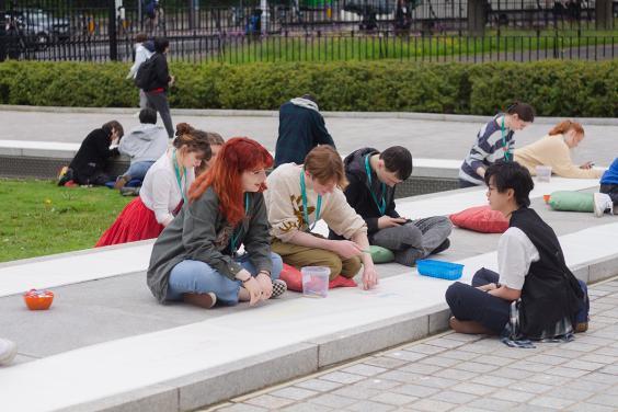 A group of young people sitting outside on stone steps, they are drawing and are surrounded by arts supplies.