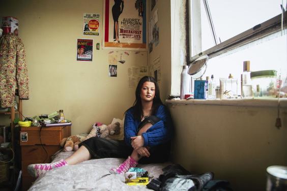 A photograph of a young woman sitting in her bedroom, surrounded by her belongings. On the wall, a number of posters and cuttings hang including a Breakfast at Tiffany's poster; on the bed itself are a few stuffed animals, and various cosmetics line the window sill.
