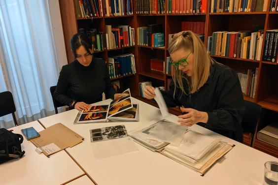 Two women sit at a desk studying documents, surrounded by books