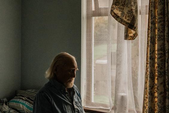 Photograph of an older man with a white beard, sitting in a small room in blue pyjamas