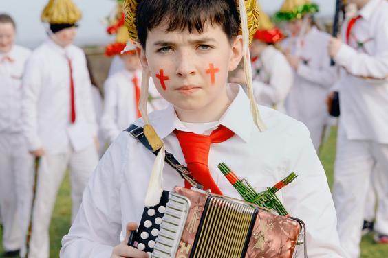 A young boy wears a traditional 'Biddy Boy' outfit and holds an accordion.