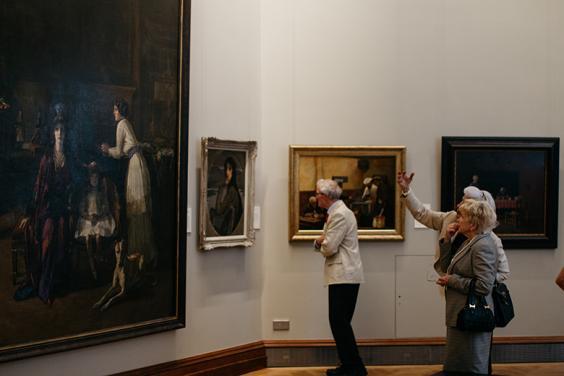 Photo of two women and a man looking at oil paintings in a gallery.