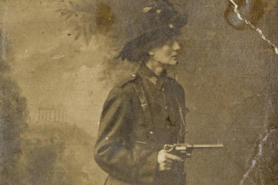 Black and white studio photograph of Constance Markievicz wearing the ICA uniform, posing against a painted backdrop of a landscape and holding a gun.