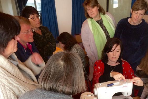 Photo of a group of women watching a seated woman use a sewing machine