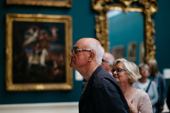 Photo of a man and woman in a gallery room with gilt-framed paintings in the background.