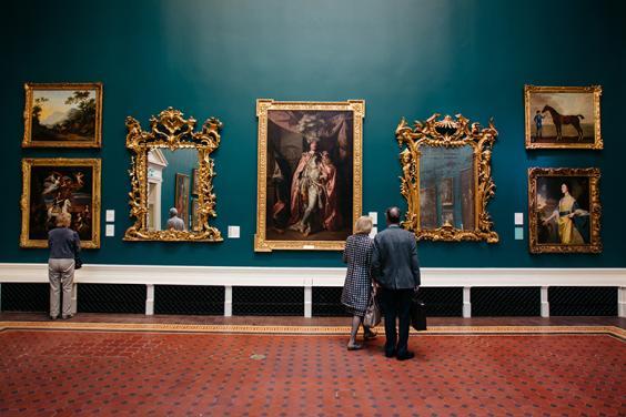 Photo of a couple standing in a gallery in front of a wall displaying gilt-framed paintings and mirrors.