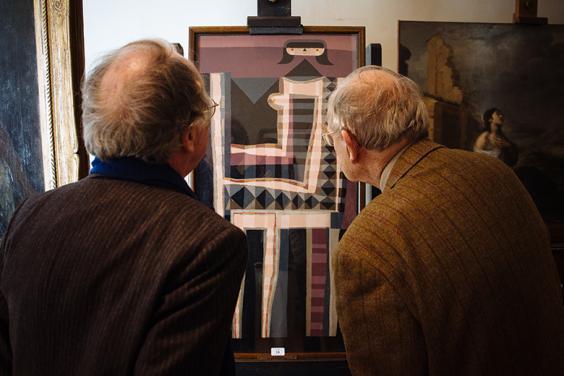 Photo of two men with their backs to the camera, looking at a painting on an easel.