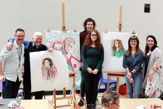 Photo of a group of people standing beside two easels holding portraits on paper.