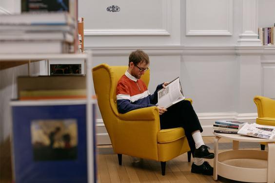 A man sits in a library on a yellow armchair reading a book