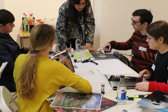 Photo of a group of young people seated at a table covered in magazines and art materials.