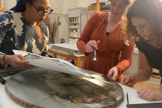 Three women looking at a painting on a table with torches and conservation equipment.