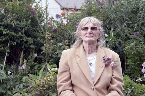 A woman in a beige coloured jacket stands in a garden, wearing dark glasses. On her lapel there is a foxglove; the garden behind is also filled with foxgloves.
