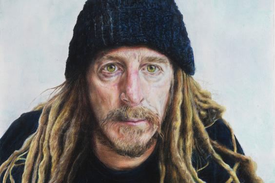 Bust-length portrait of a man with a short beard, blond dreadlocks and wearing a dark woolen hat