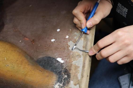Close-up view of two hands holding tools against a damaged area of an oil painting